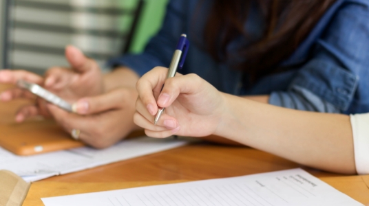 A hand holds a pen, with another person in the background on a cell phone